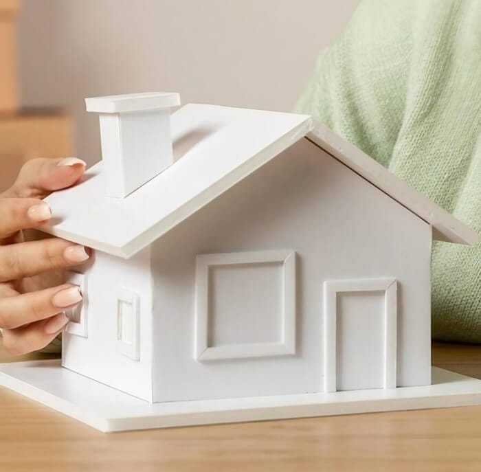 A person holding a small white model house with detailed features, resting on a wooden table, with a soft-focus background.